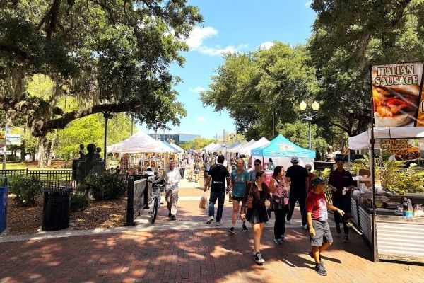 Crowds walk along a pathway lined in event tents with trees hanging overhead at Fall Fiesta in the Park at Lake Eola.