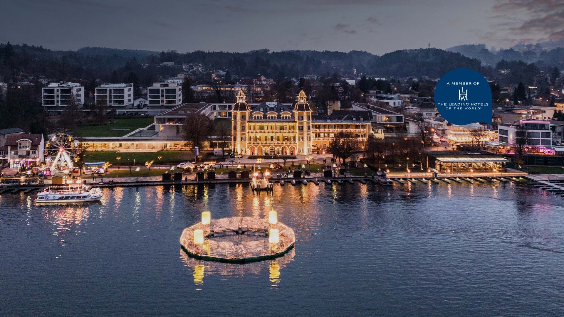 Illuminated waterfront hotel with floating circle lights and illuminated Ferris wheel at Falkensteiner Schlosshotel Velden