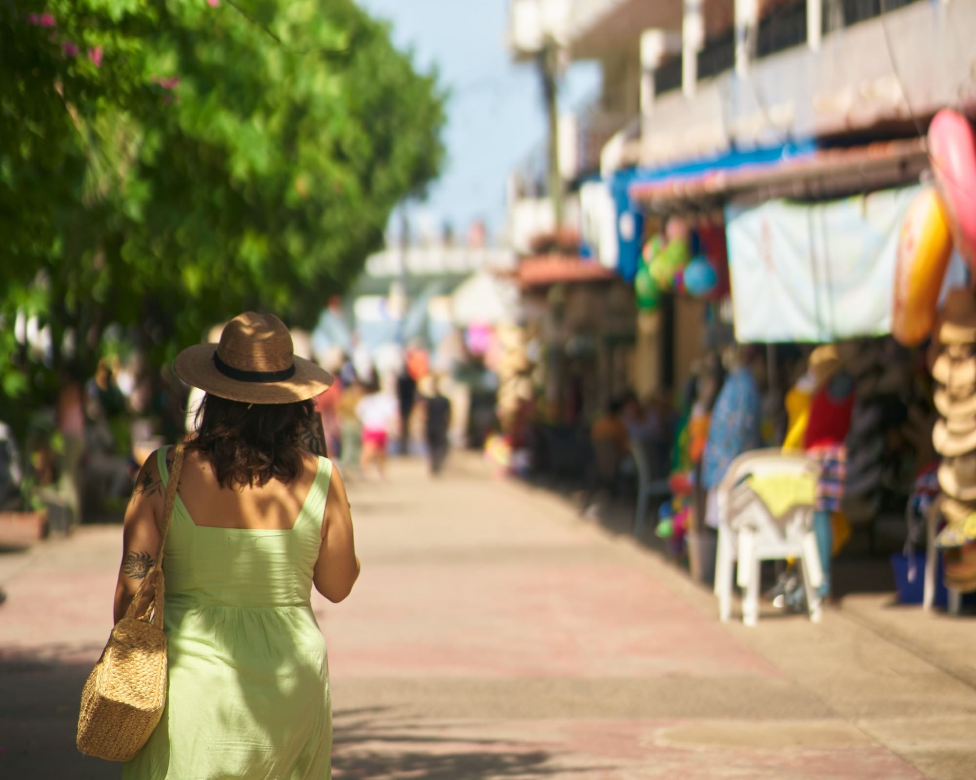 A woman wearing a straw hat and a light green summer dress walking through a sunlit outdoor market lined with local shops and lush green trees.