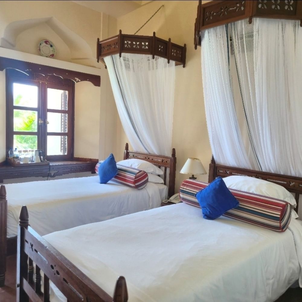 Two beds with mosquito nets and blue cushions in a Standard Twin Room at Zanzibar Serena Hotel.