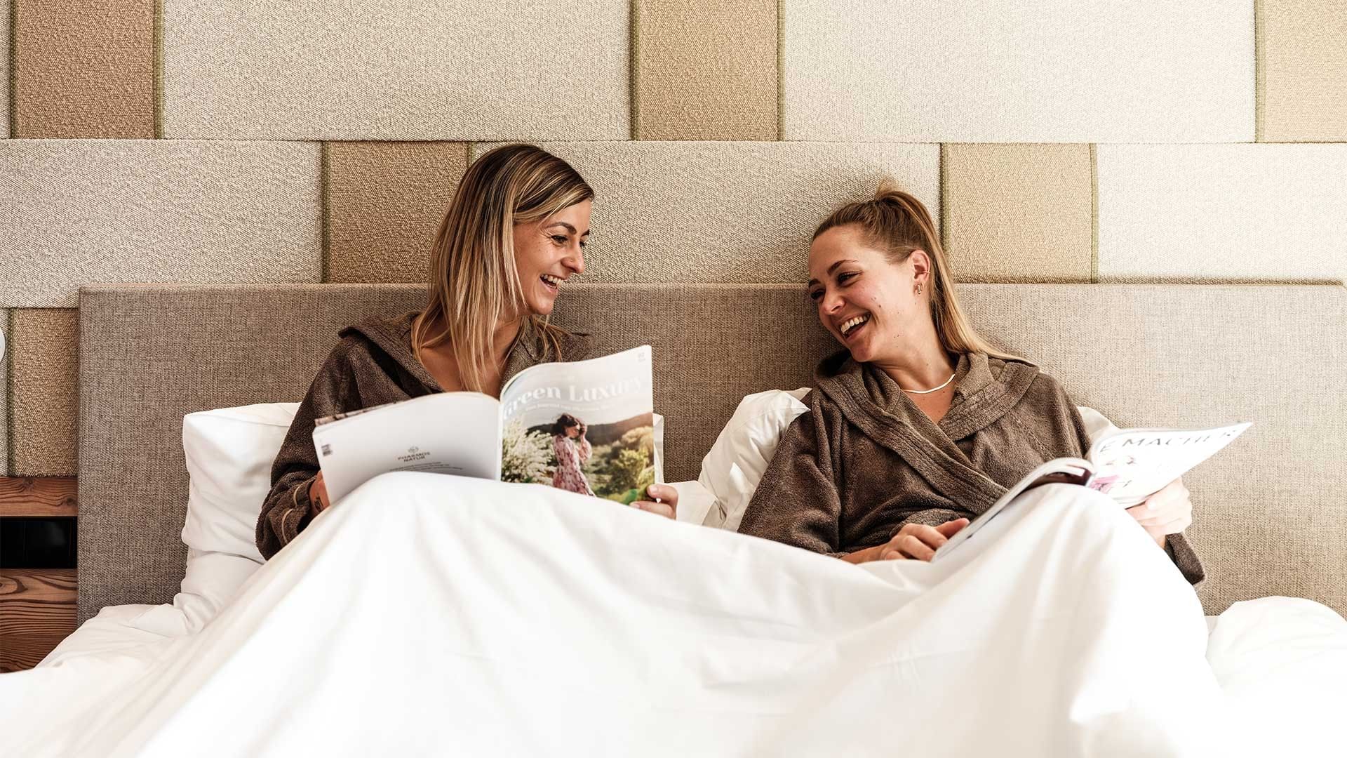 Zwei Frauen in Bademänteln liegen im Bett und lesen Zeitschriften.