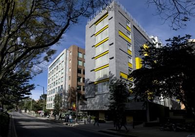 Edificio gris con ventanas amarillas en una calle con árboles y peatones.