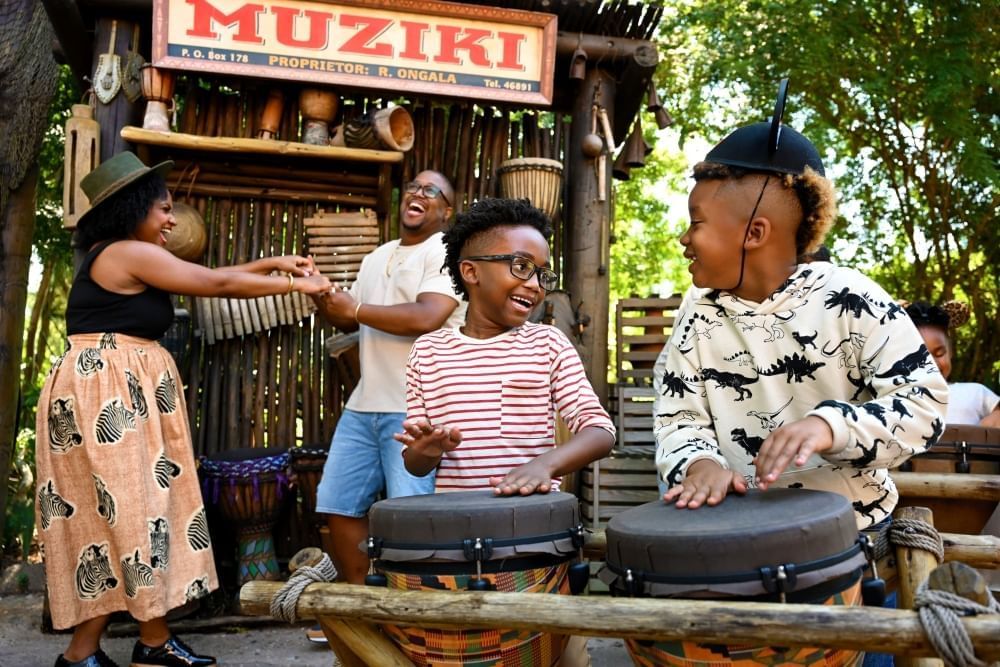 Two boys, one in Mickey ears, smile while playing African drums as a man and a woman dance in the background.