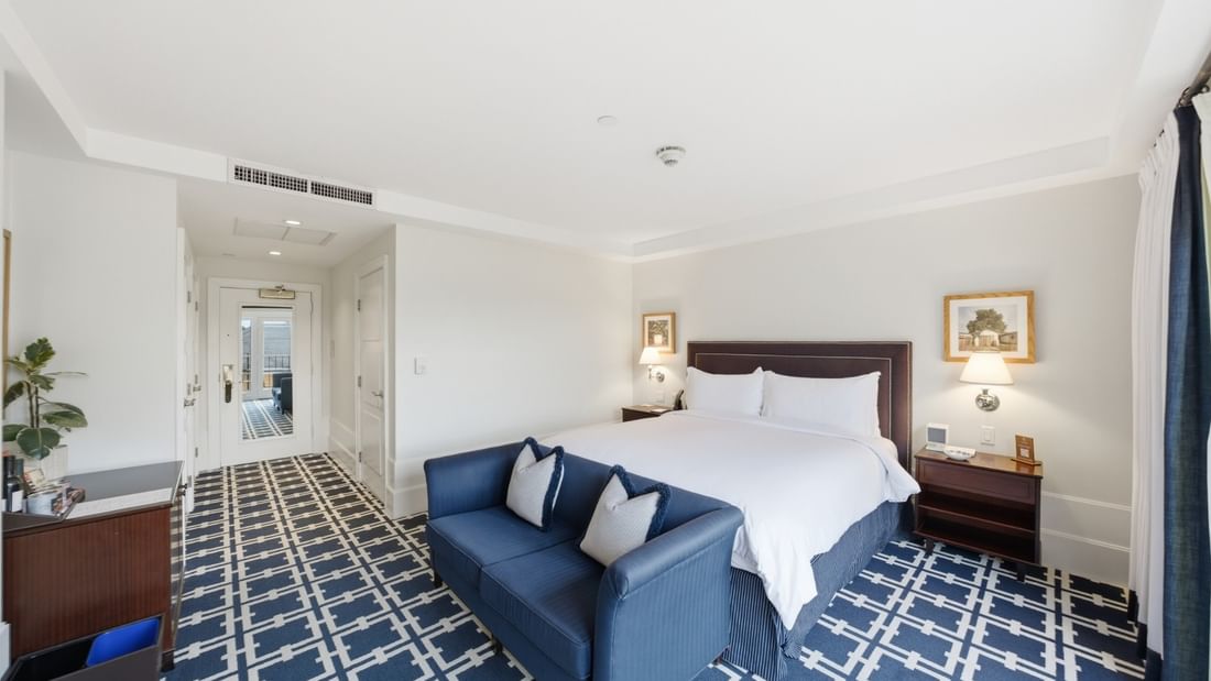 Deluxe King room with white bedding, blue sofa, patterned carpet, and wall sconces at el PRADO Hotel, Palo Alto.