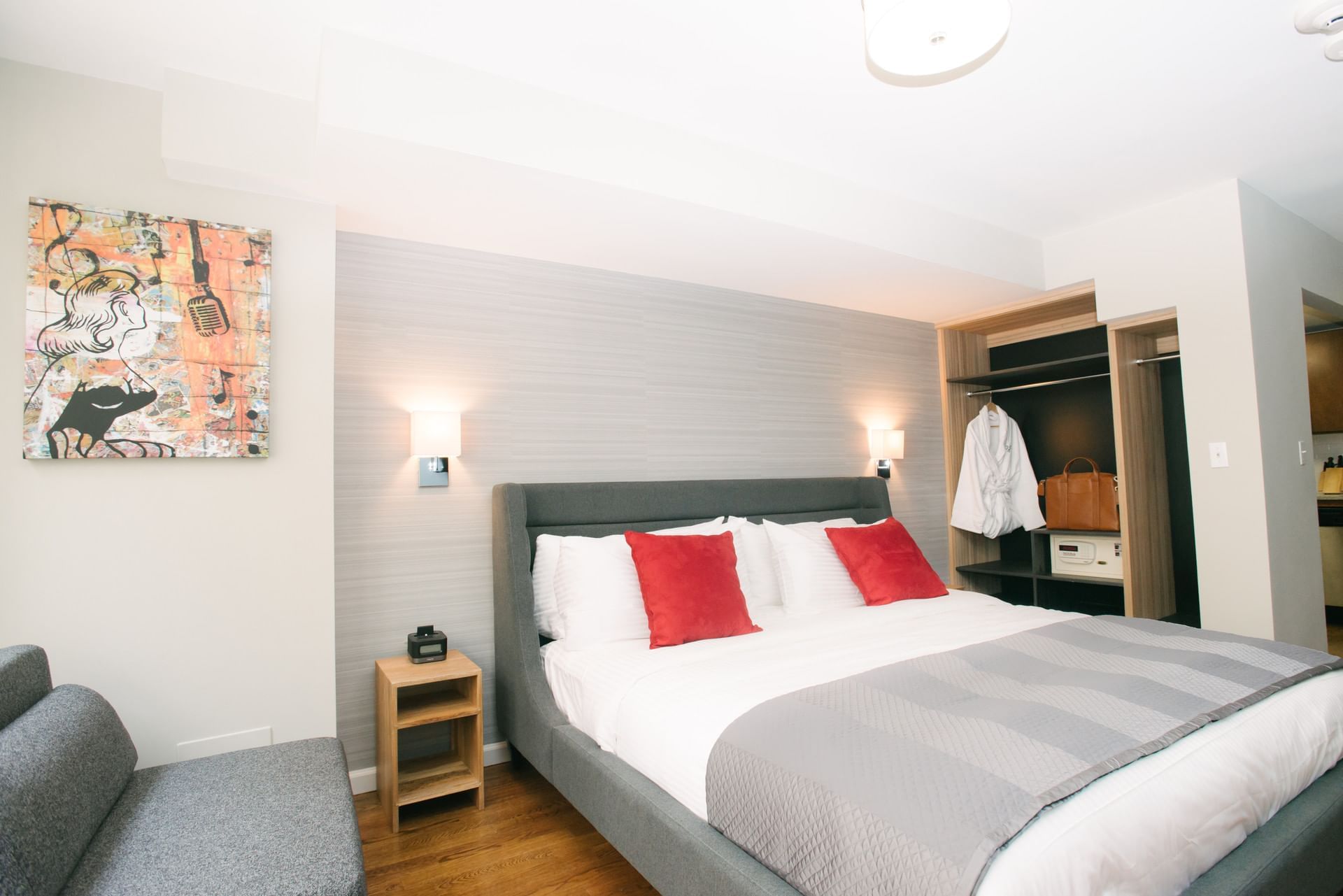 A hotel room with a king-sized bed, gray upholstered headboard, white bedding, red pillows, a gray sofa, wooden nightstand, and closet with a safe and white robe.