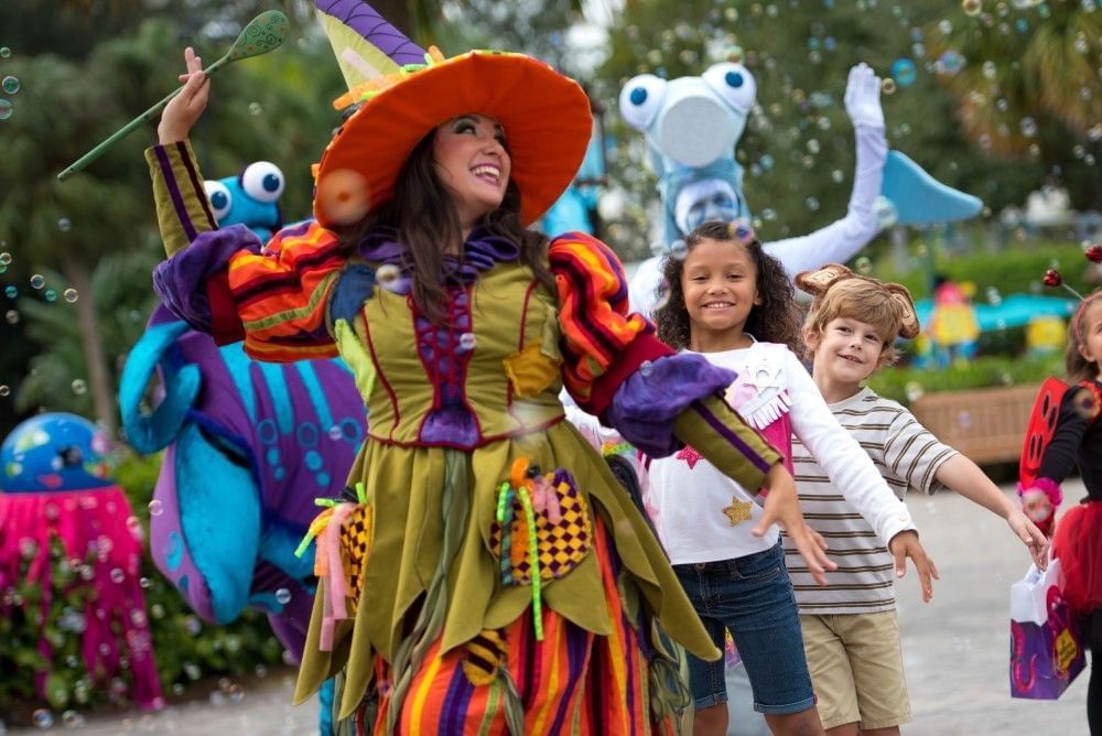 A woman dressed as a colorful witch leads a parade of children outside.