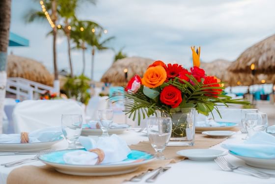 Beachside table set for dinner with flowers, plates, and beach huts in the background.