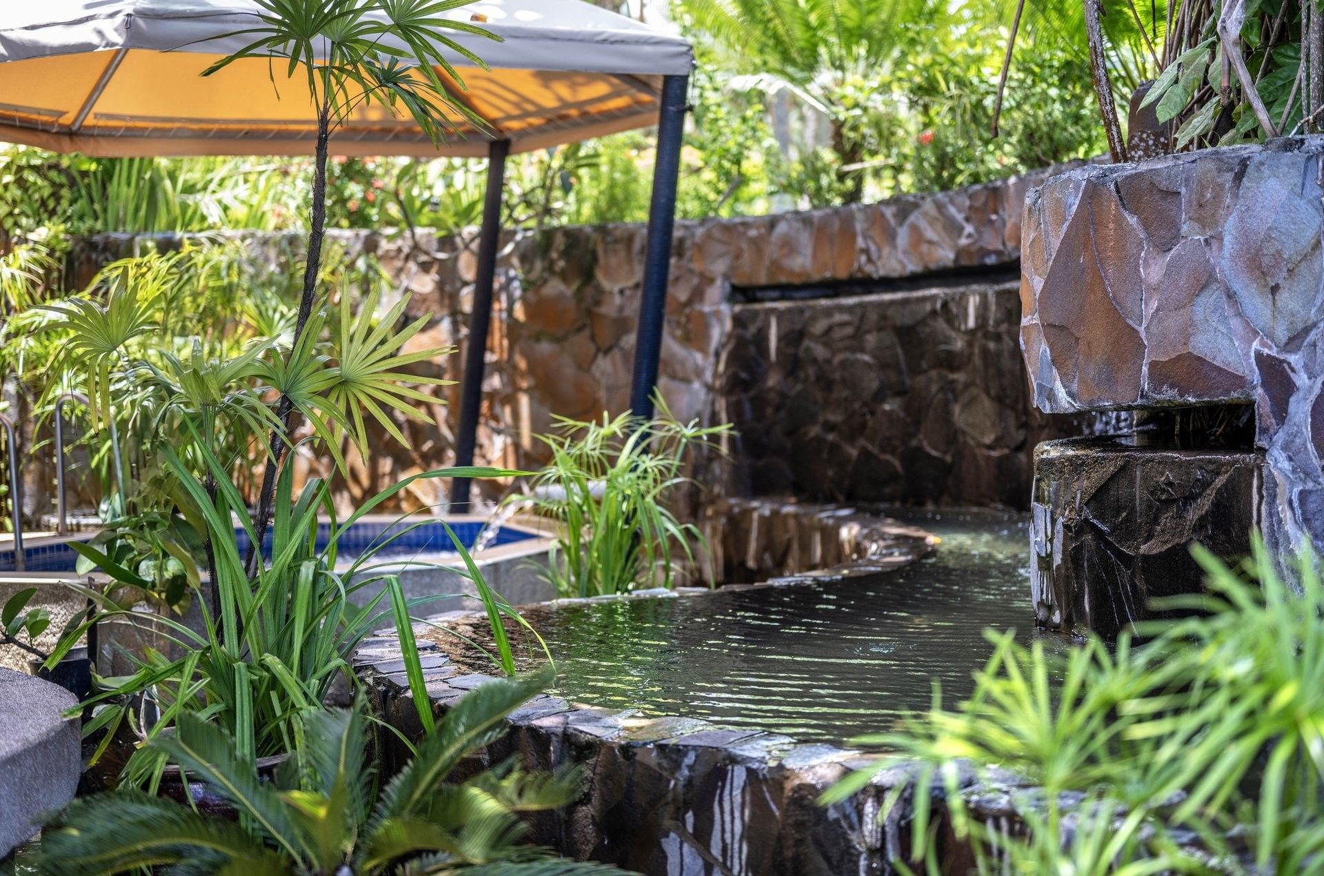 Stone waterfall flowing into a dark pool by green ferns under a tan gazebo at Warwick Fiji Resort and Spa