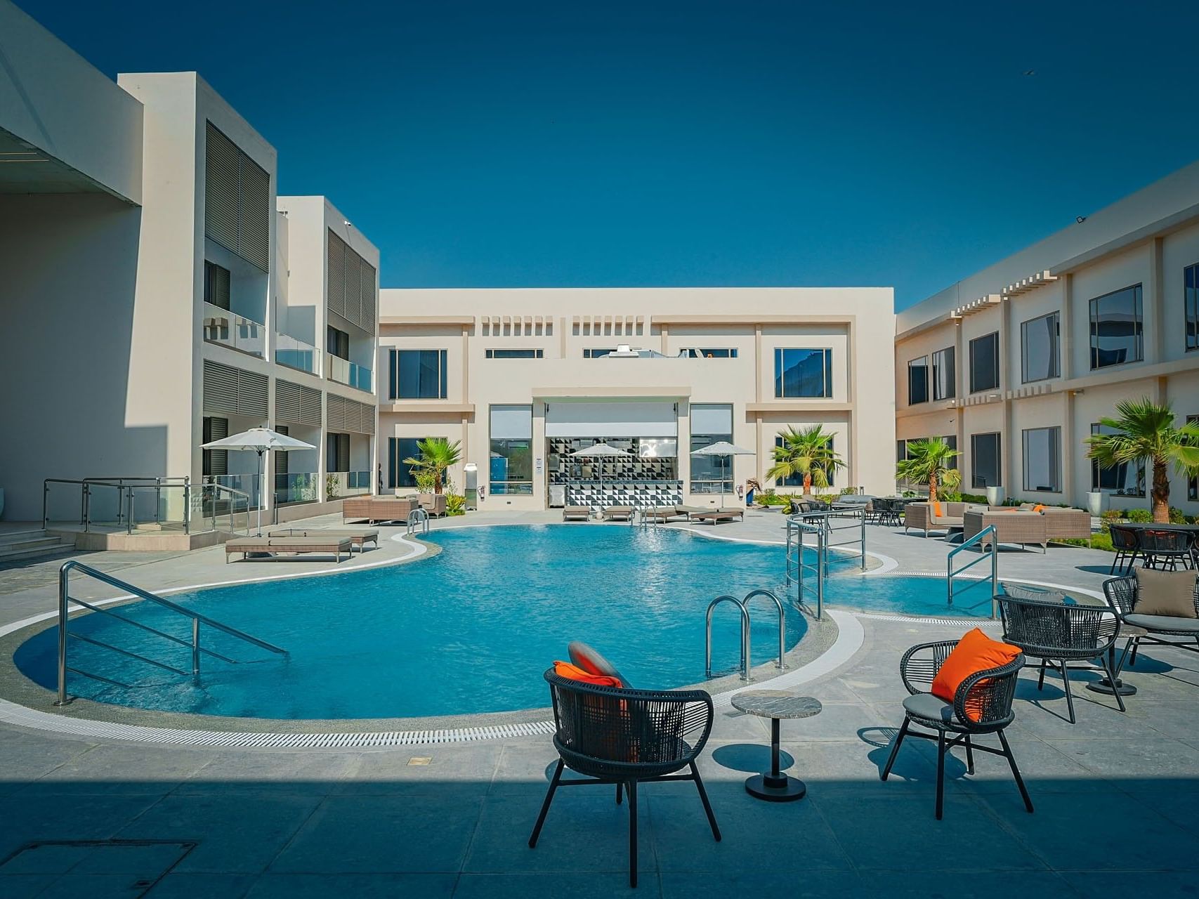 Outdoor pool area with lounge chairs, tables, and modern buildings under a clear blue sky.
