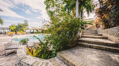 Curved stone steps with tropical plants lead to a swimming pool area under a bright sky at Hotel Montana Haiti