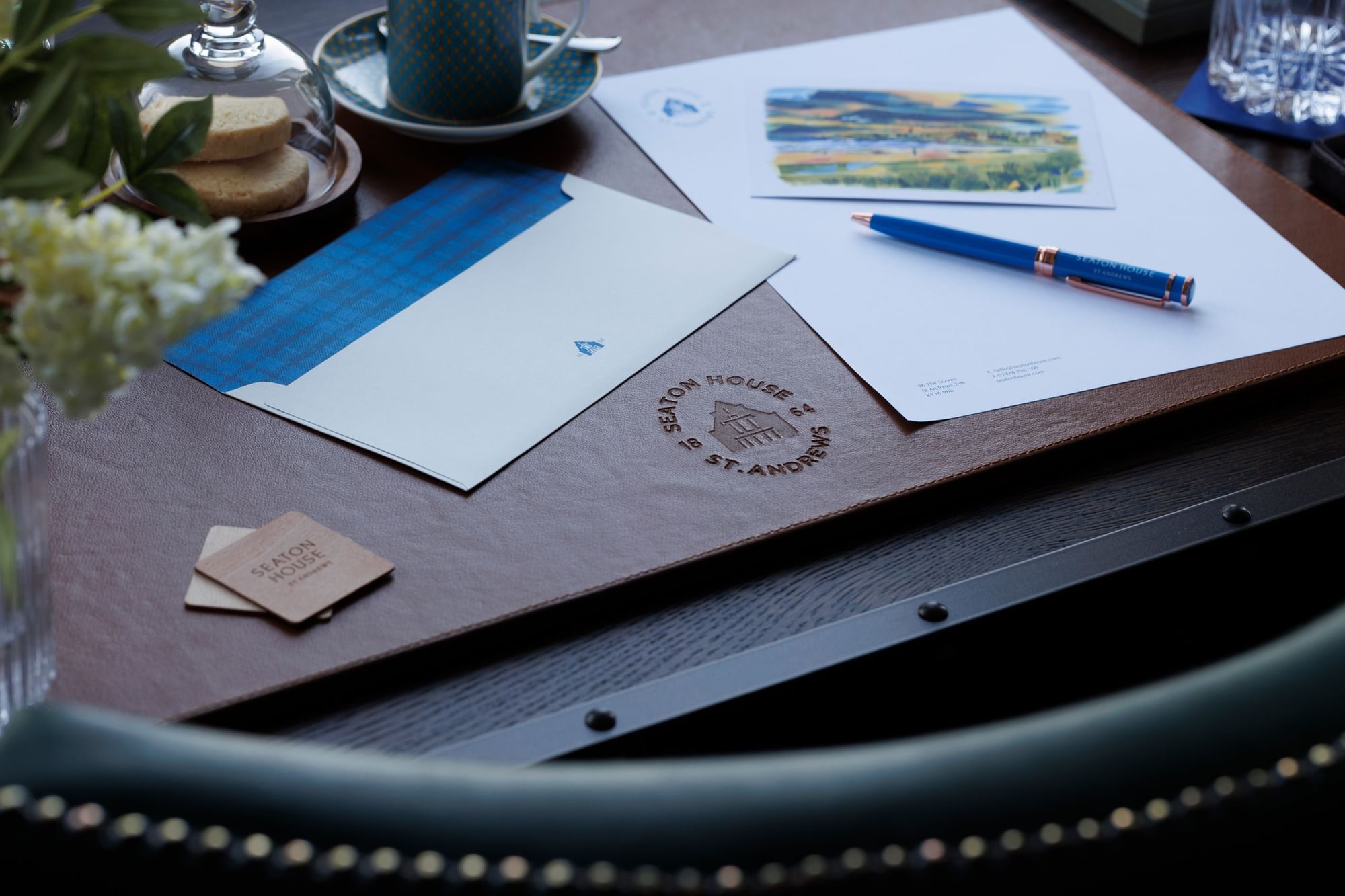Branded notepad and pen on a leather desk blotter, with a teacup and a small plate of cookies at Seaton House