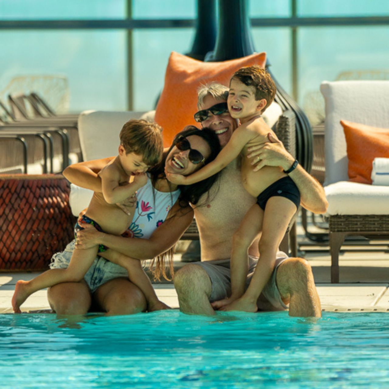 Happy family with two young children hugging in swimming pool beside lounge chairs and orange cushions at Hotel X Toronto