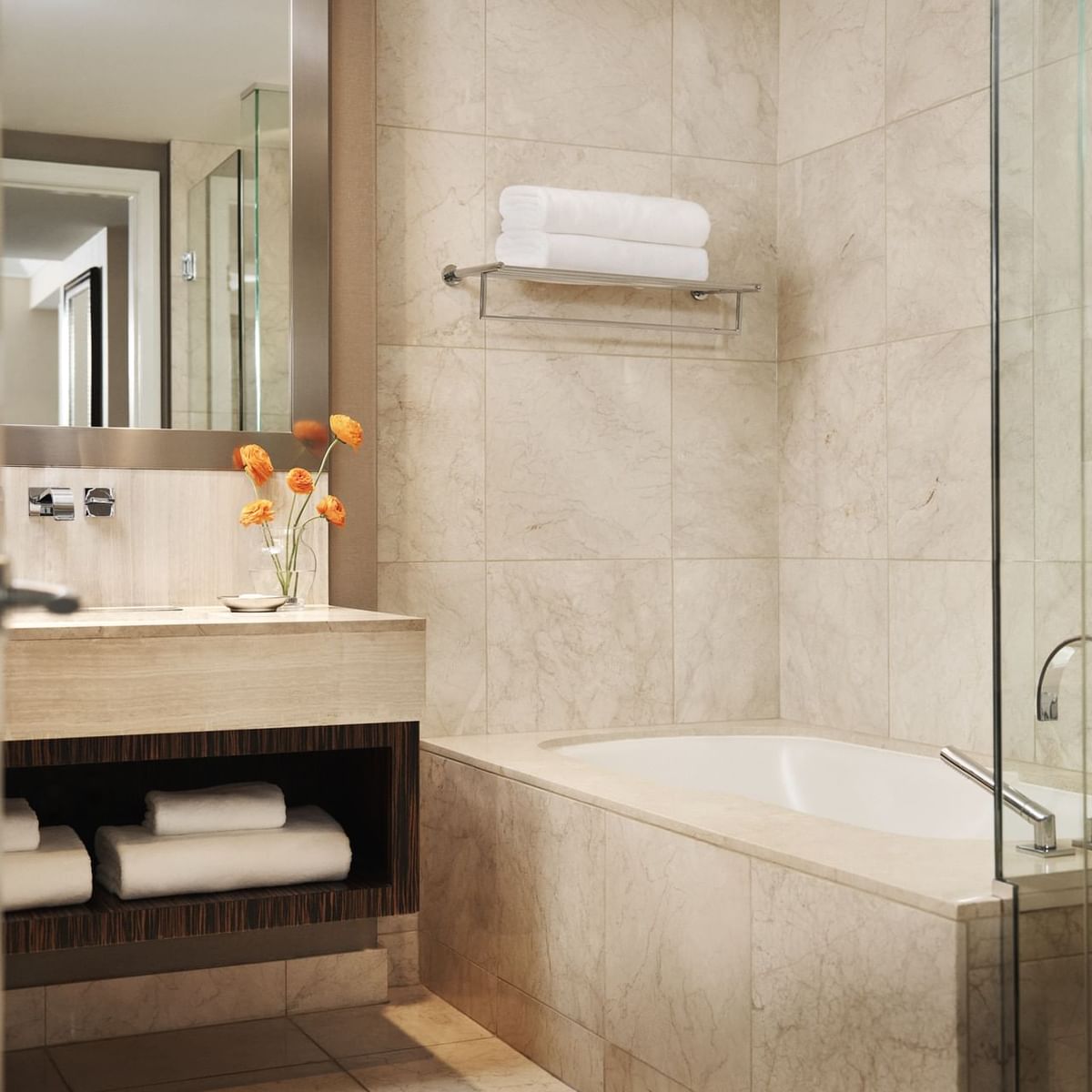 A bathroom with a bathtub, towel rack, towel, mirror, sink, and orange flowers in a vase.