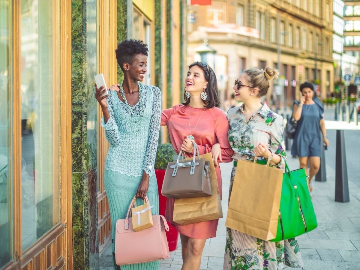 three woman walking down a city street holding shopping bags looking at each other laughing