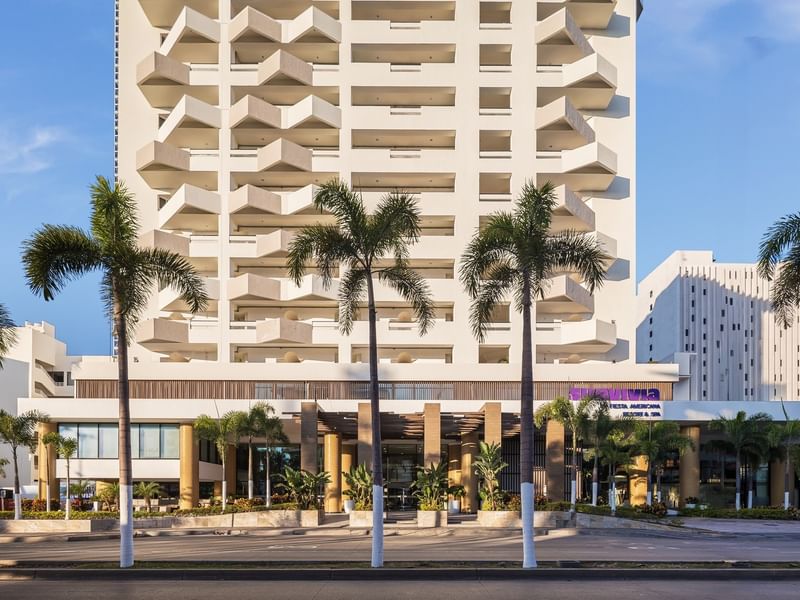 Front facade of a tall white Sunvivia Mazatlán resort building with unique geometric balconies and palm trees along the road