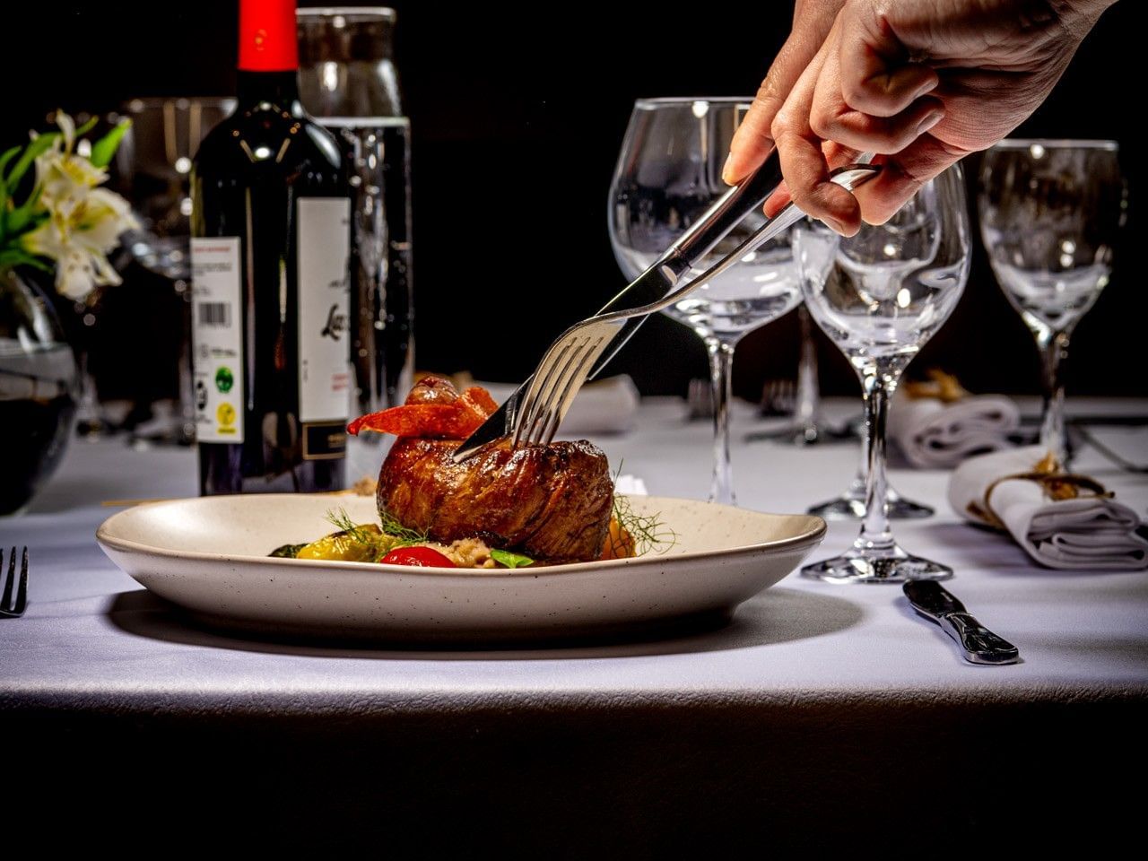 Mano tatuada sosteniendo un tenedor sobre un plato de carne, junto a copas de vino y una botella.