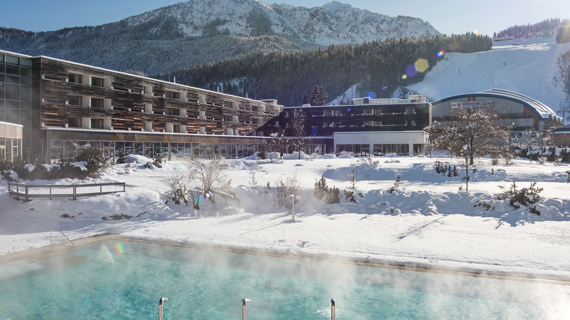 Snowy scene with a steaming pool in the foreground and a mountainous backdrop at Falkensteiner Hotel and Spa Carinzia