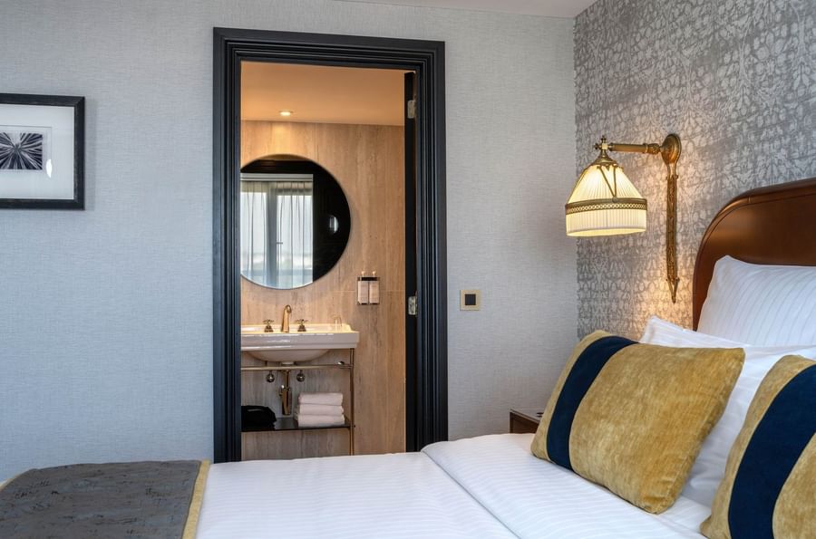 Yellow pillow on bed by lamp, with white sink and round mirror in the bathroom in Warwick Suite at Hotel Barsey by Warwick