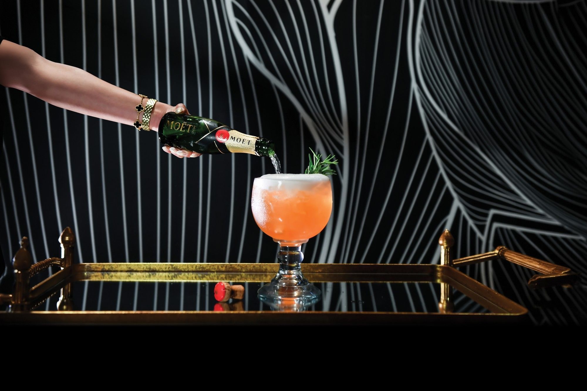 Champagne pouring into orange cocktail on gold tray against black and white striped background wall at The Brook