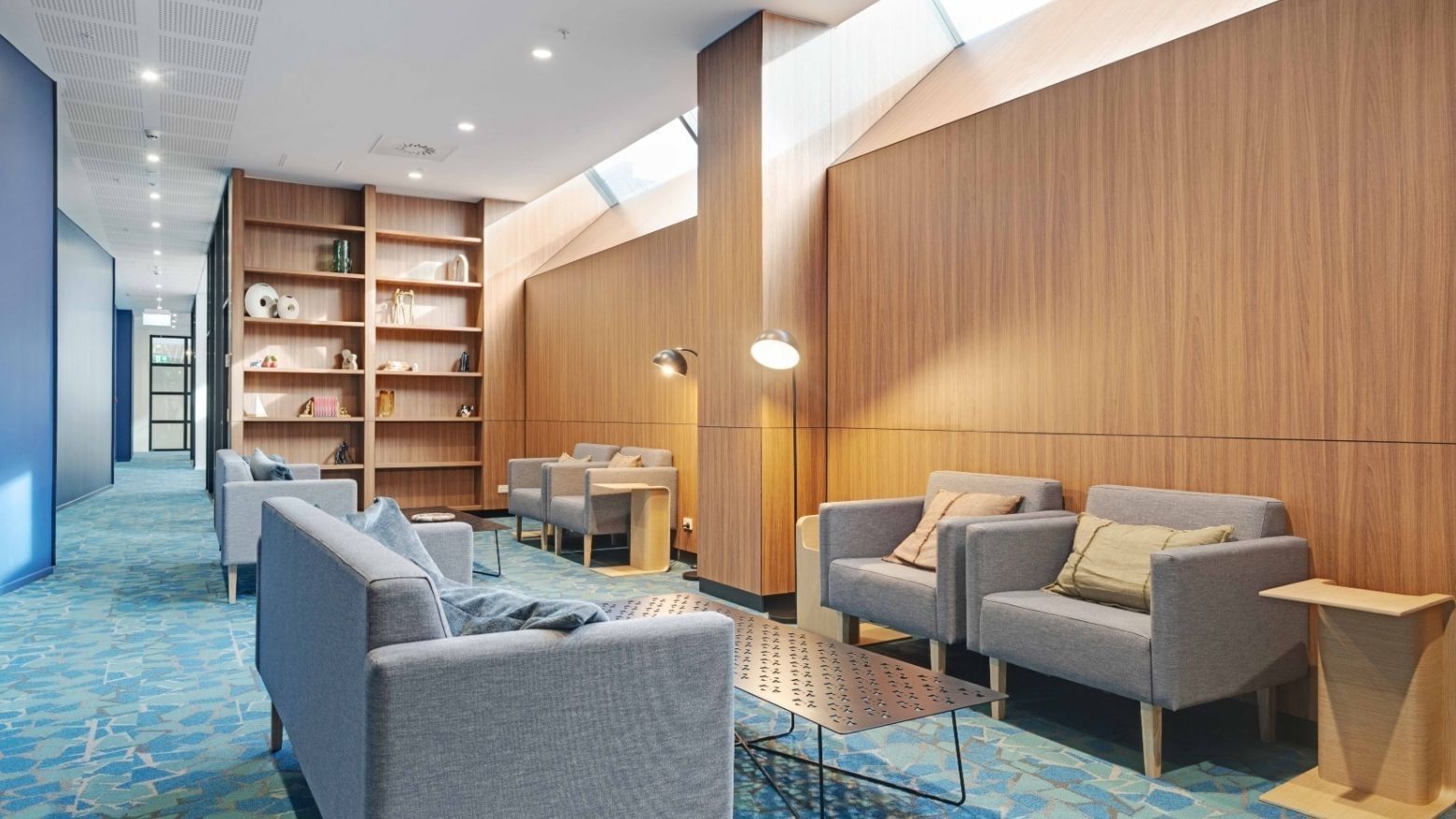 Modern communal lounge with gray sofas, wooden shelving, skylights, and blue patterned carpet at UniLodge Auckland Central
