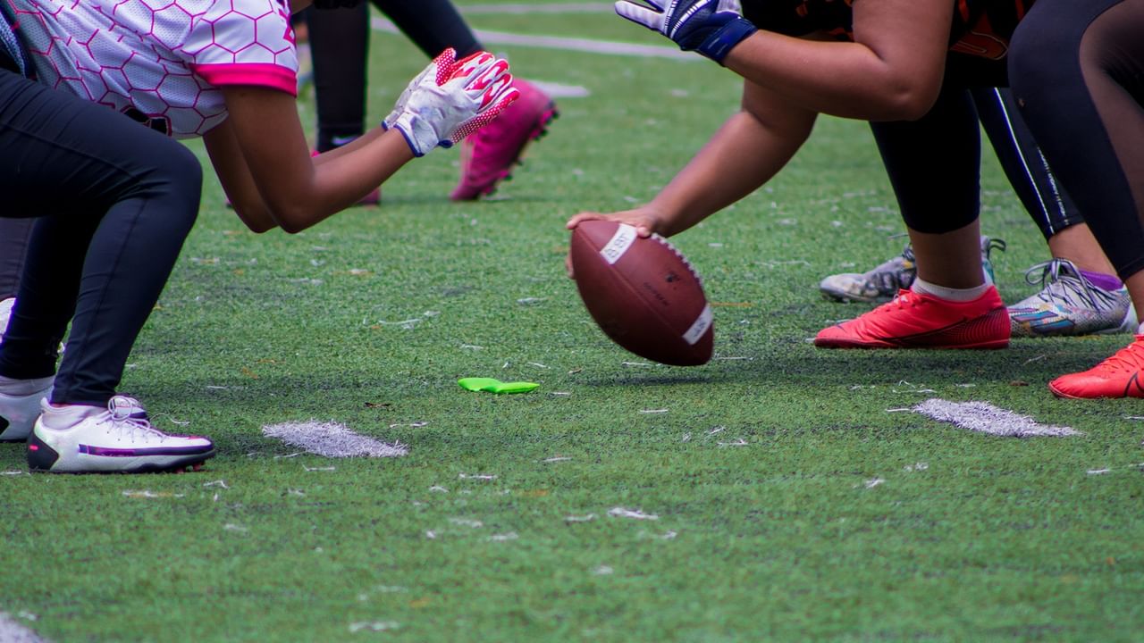 Football players in colorful cleats and gloves positioned at the line of scrimmage ready for the snap on a grass field.