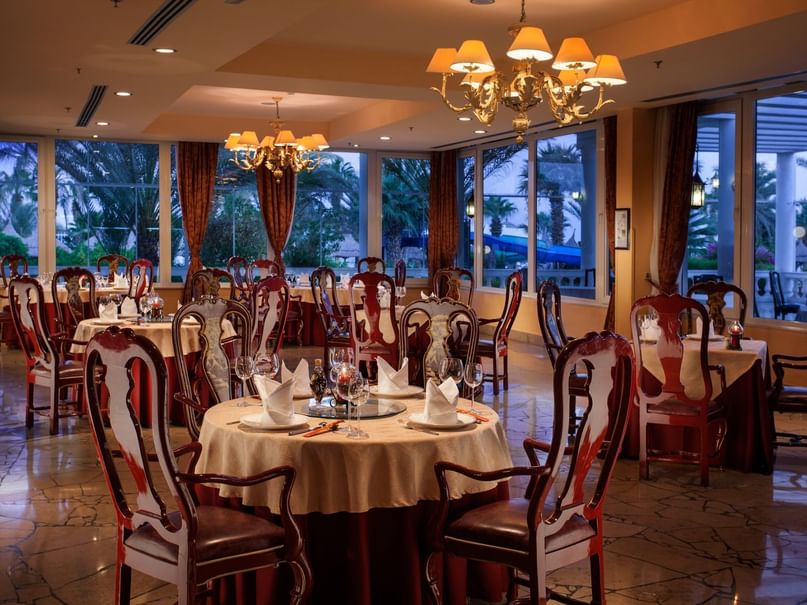 Elegant restaurant with large windows, chandeliers, and neatly set round tables with chairs.