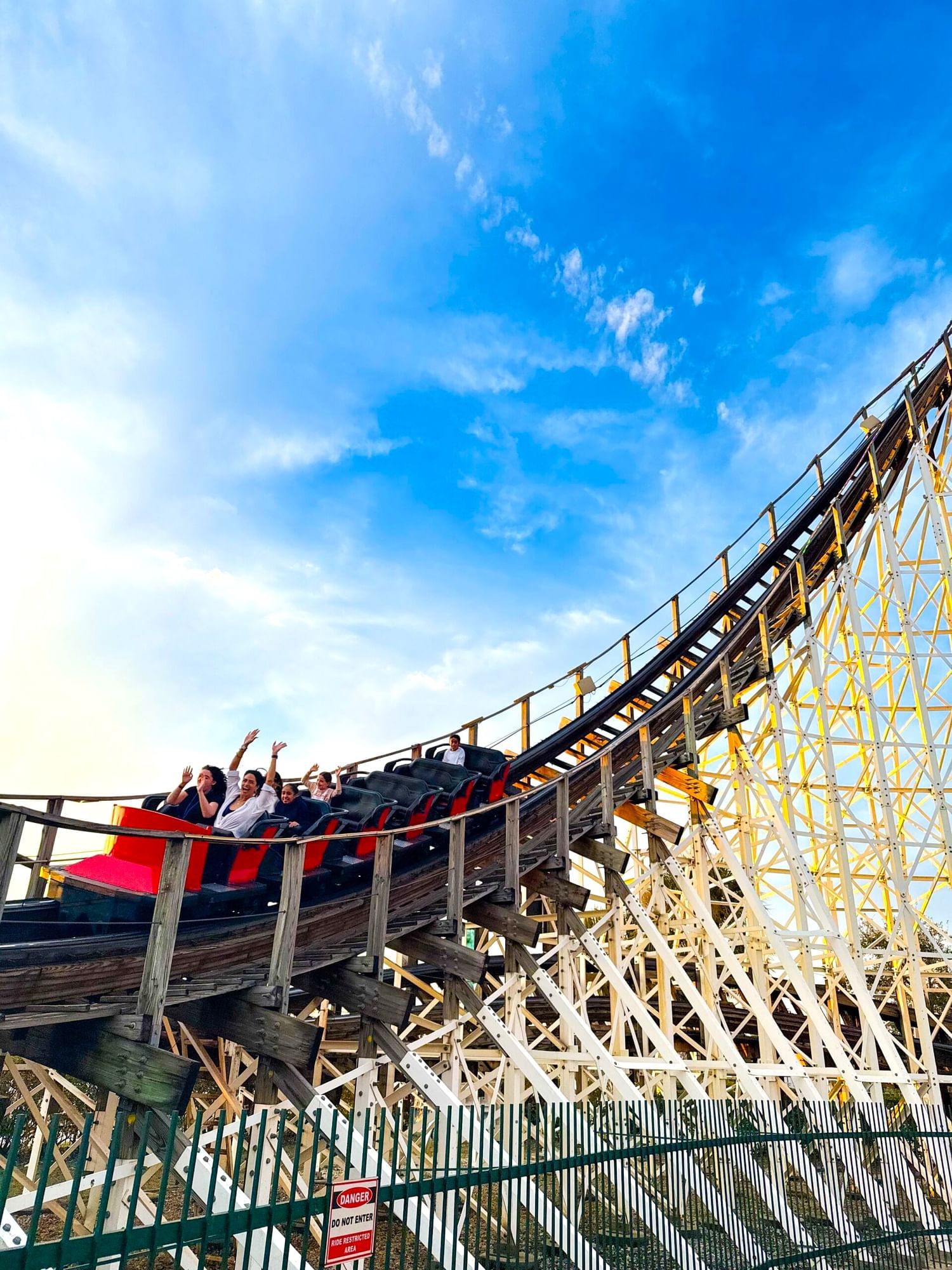 Passengers excitedly glide down a wooden roller coaster against a blue sky.