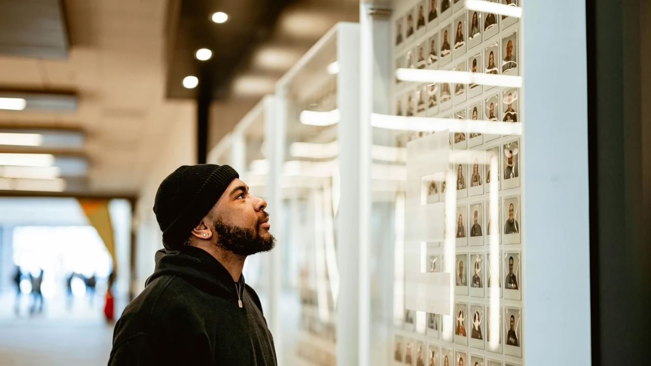 A man in a black beanie and hoodie attentively viewing a wall display of portraits inside an art gallery.