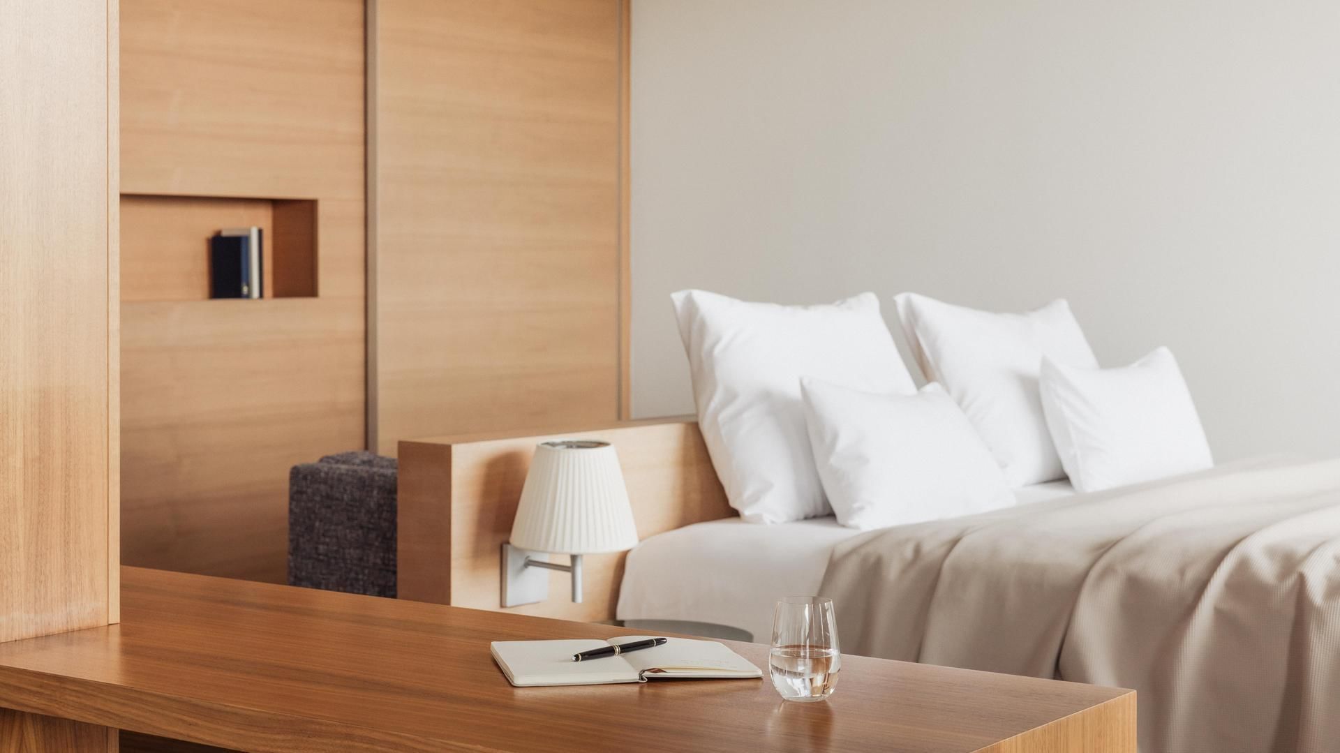 Elegantes Hotelzimmer mit Bett und weißen Kissen, Schreibtisch mit Buch und Glas Wasser.