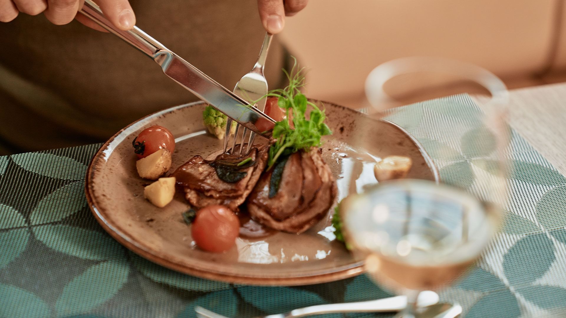 Person schneidet ein Teller mit Fleisch, Kartoffeln, Tomaten und Gemüse mit Messer und Gabel.
