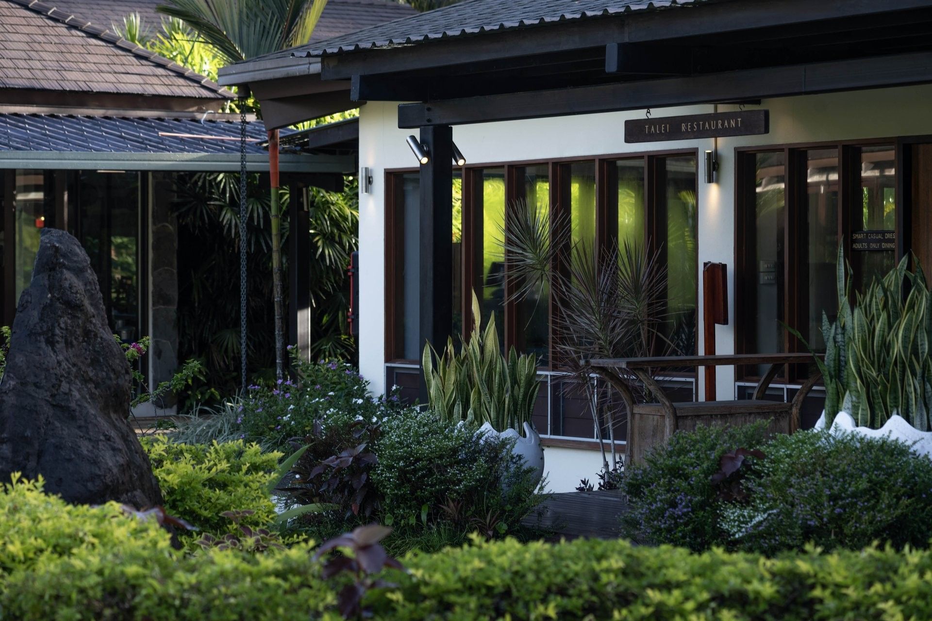Extirior view of Talei Restaurant by glass windows under a dark roof near tropical shrubs at The Naviti Resort - Fiji