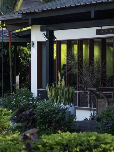 Exterior view of Talei Restaurant by glass windows under a dark roof near tropical shrubs at The Naviti Resort - Fiji