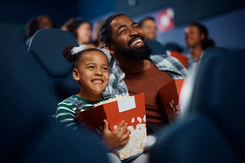 A girl in a movie theatre holding a large bucket of popcorn smiles up at a movie screen next to her dad, who is also laughing. 