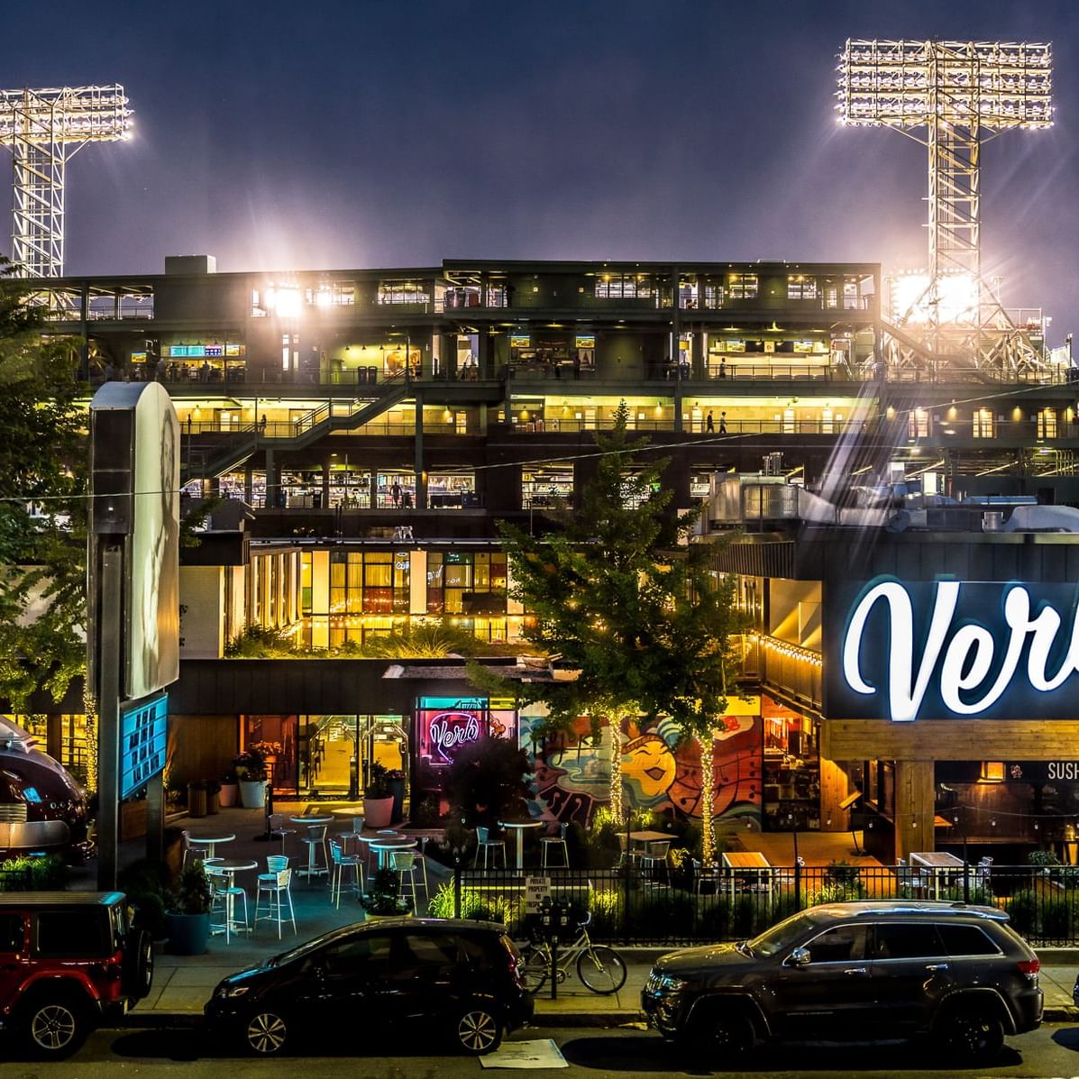 Illuminated Fenway Park stadium towers over nearby restaurants and shops during evening twilight in Boston's Kenmore Square.