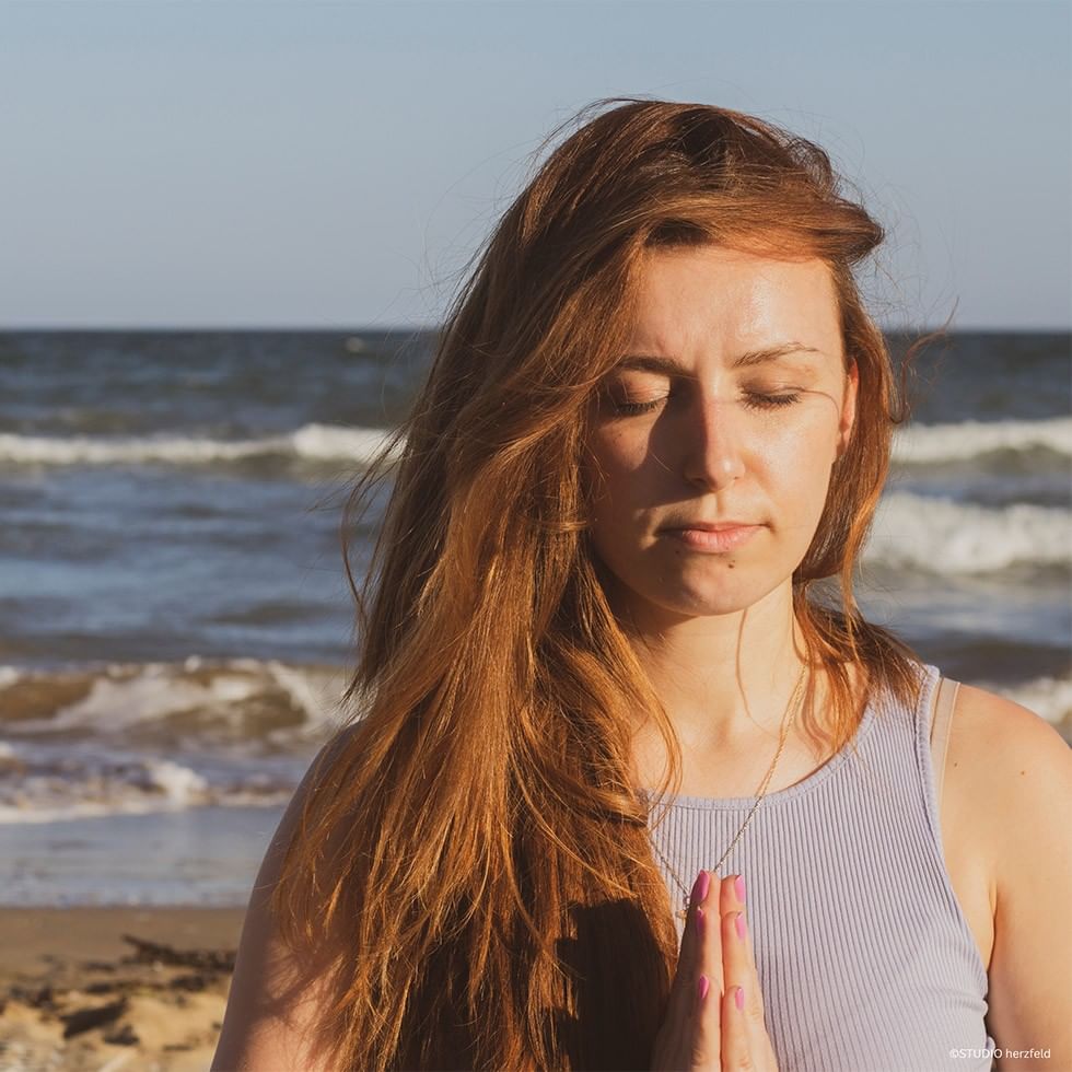 Woman meditating by the ocean with her hands in prayer position at Yoga Experience with STUDIO herzfeld.