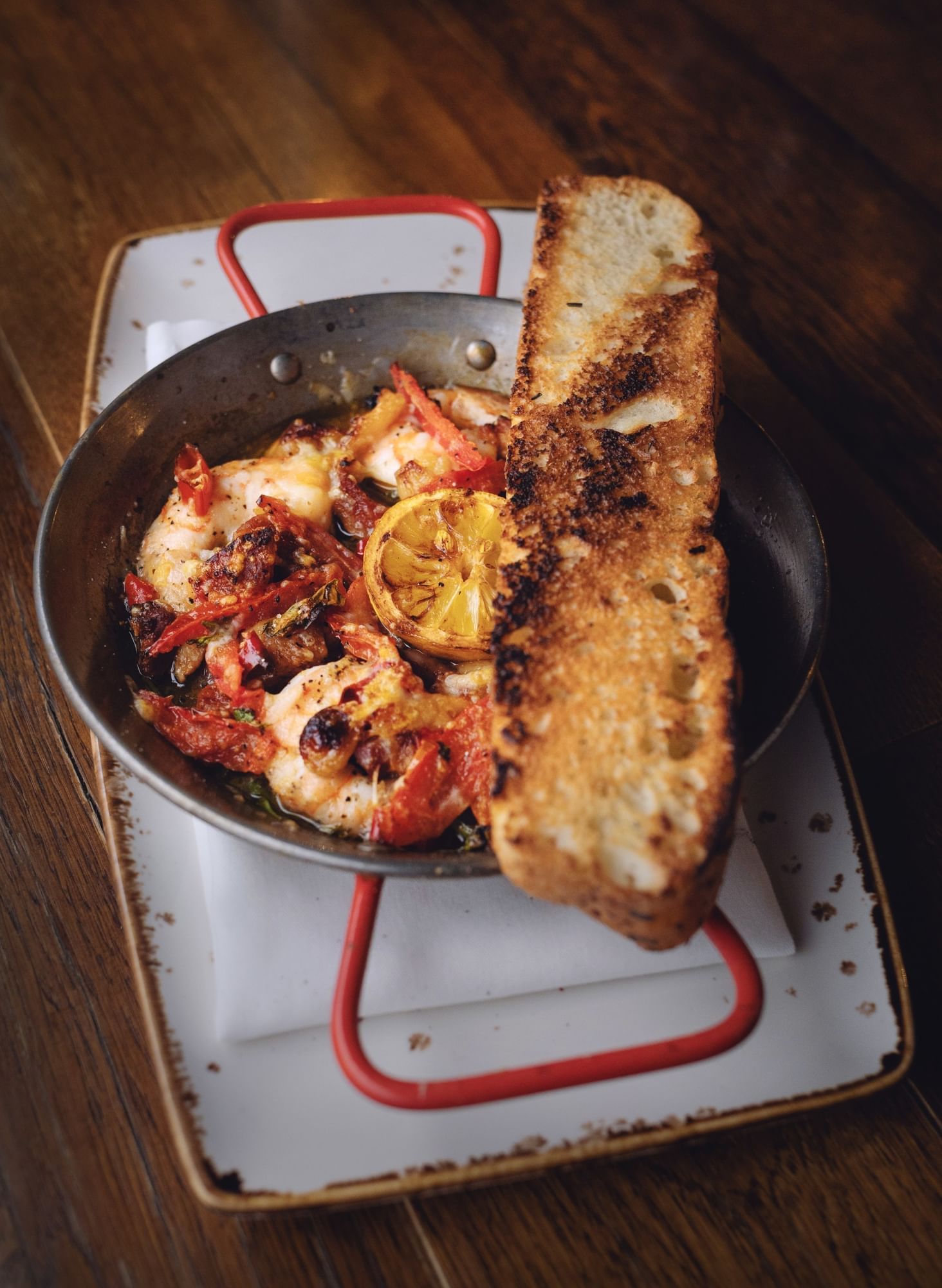 A small pan with shrimp and tomatoes with a round lemon slice on top and toasted slice of bread