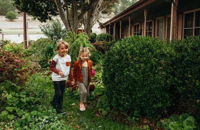 Two children holding hands, walking through a lush garden near lodge