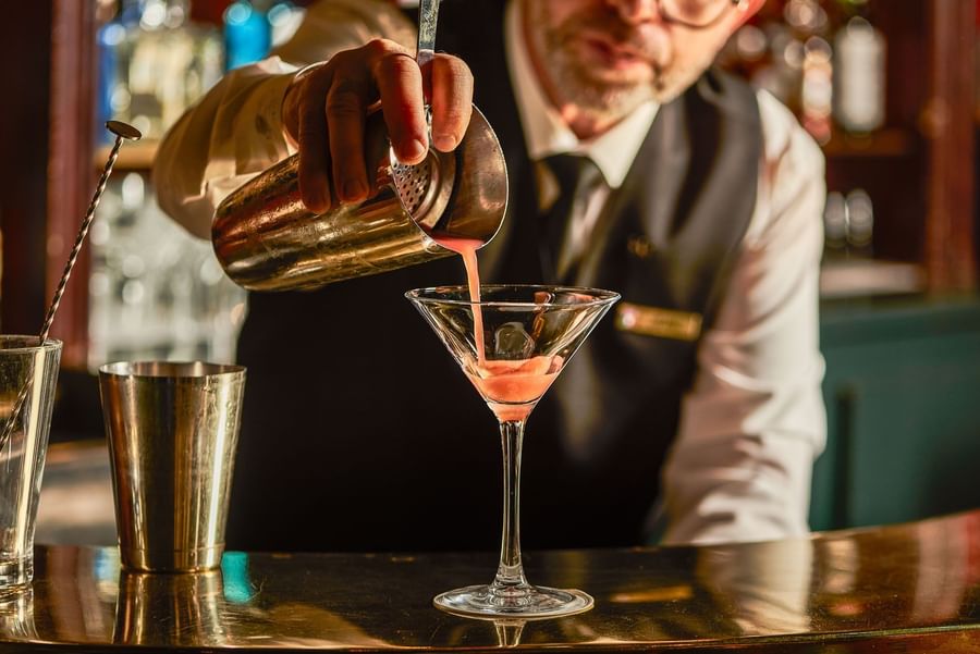 Bartender pouring a pink drink into a glass with a metal shaker on a bar counter at Hotel Westminster Paris