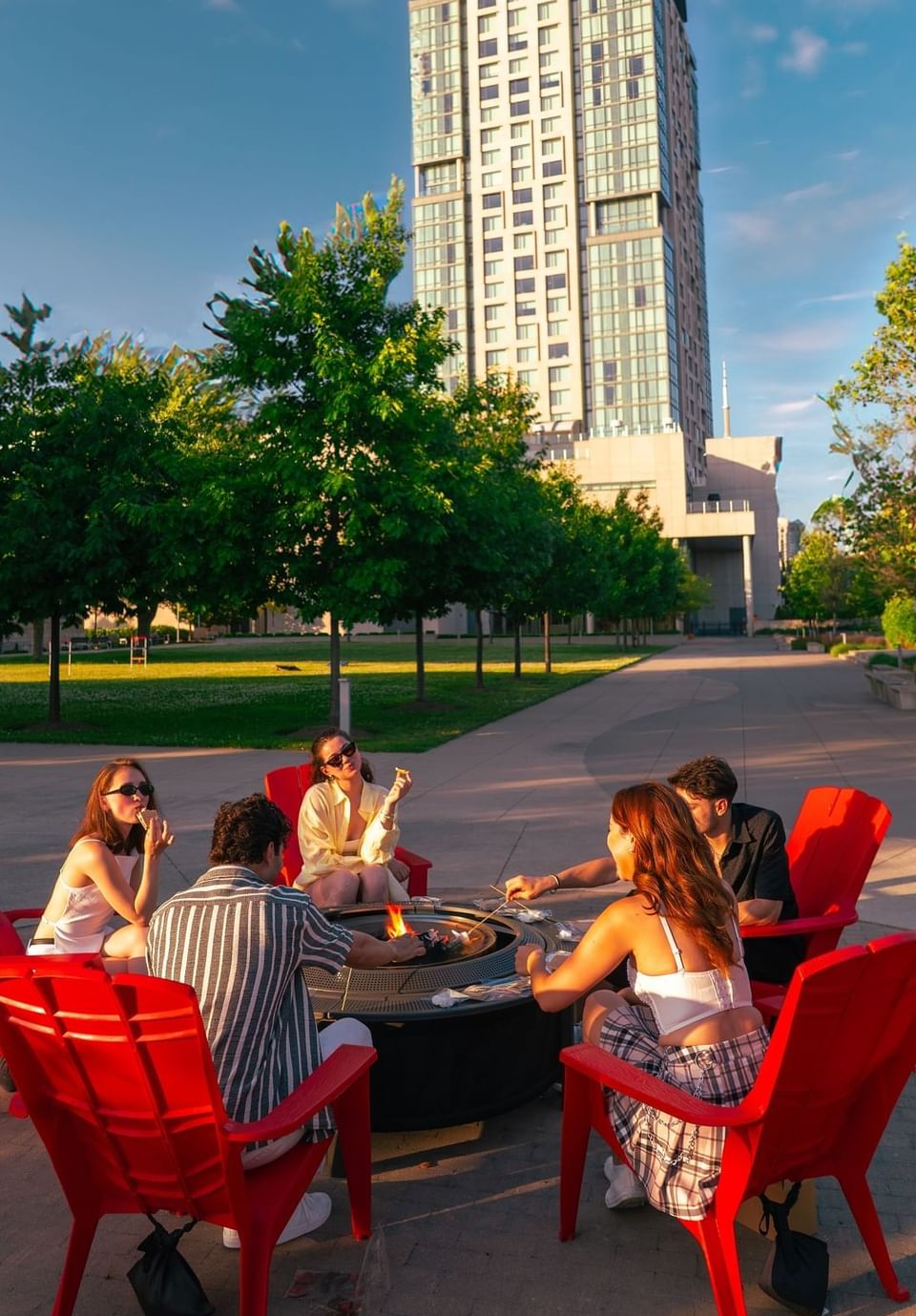 Group of young adults sitting in red chairs around a burning fire pit on a large paved courtyard at Hotel X Toronto