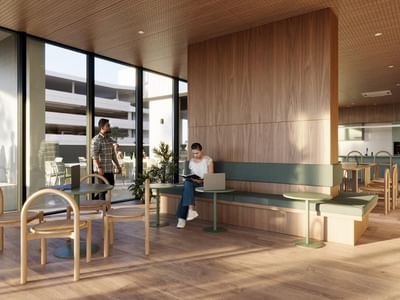 A modern lounge area with wooden tables, chairs, and a green bench, featuring large windows and an outdoor view.