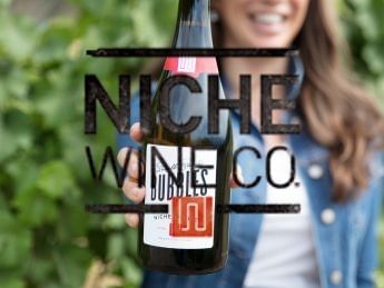 Smiling woman holding Niche Wine Co. bottle outdoors.