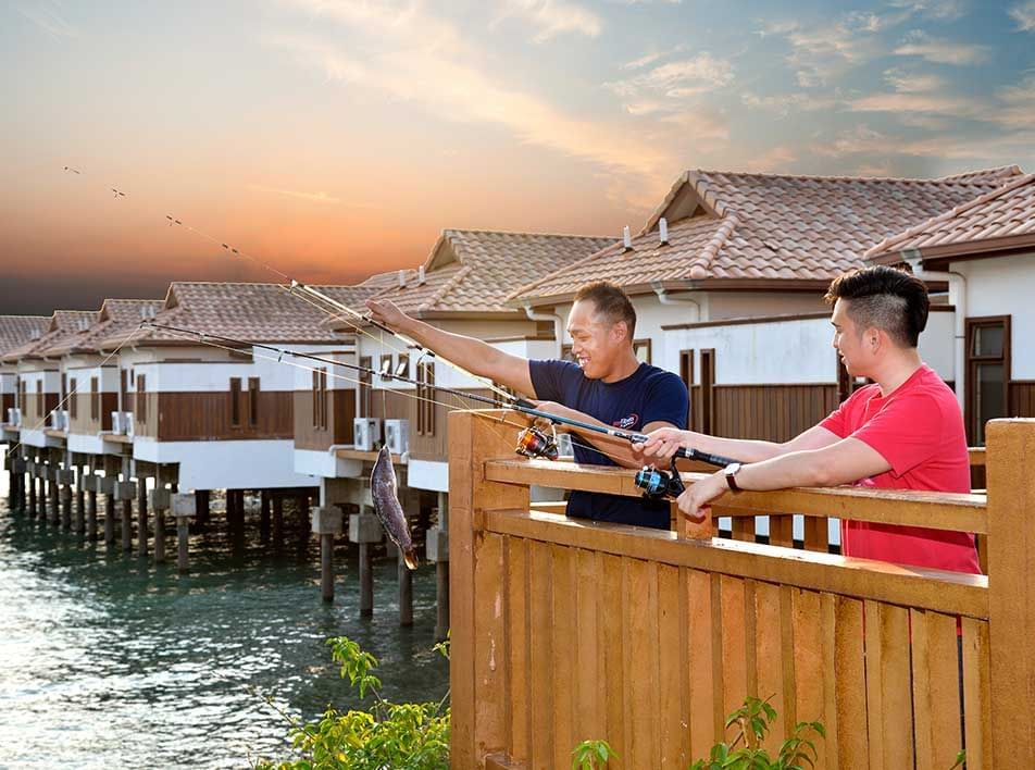 Two people fishing off a pier at sunset with stilt houses in the background - Lexis Port Dickson