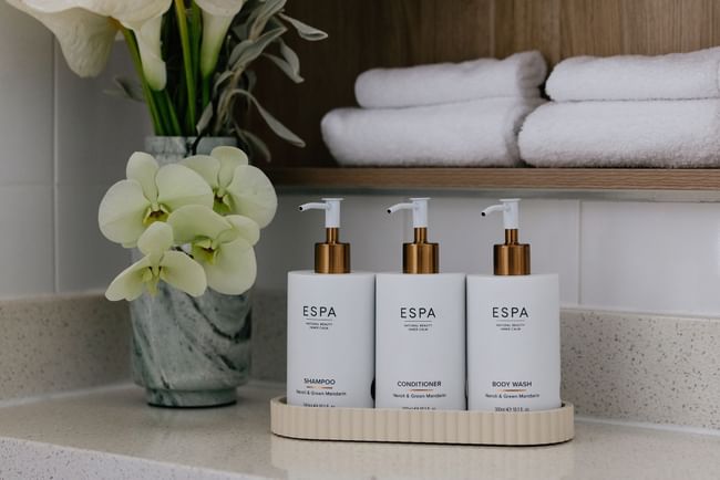 Elysium Manly bathroom counter featuring three luxury amenity pump bottles placed neatly beside a vase of fresh orchids