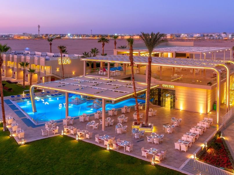 Aerial view of a resort with pool, palm trees, and tables at dusk, surrounded by desert landscape.