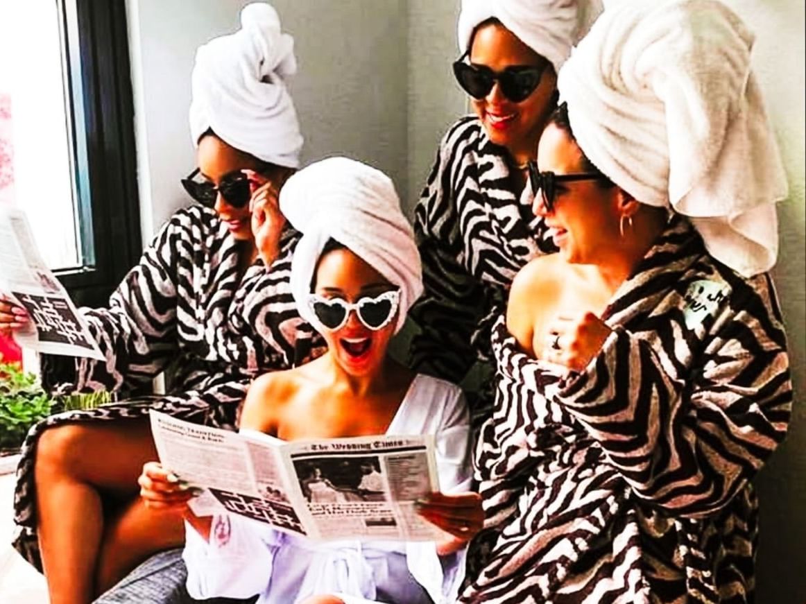 Group of women in matching zebra-print robes, white towel wraps, and sunglasses reading newspapers during The Basic B.