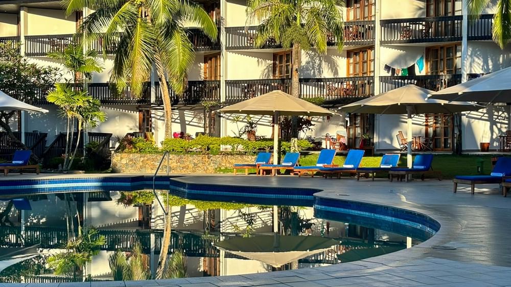 Blue swimming pool with beach chairs under umbrellas in front of resort building at Warwick Fiji Resort and Spa Korolevu.