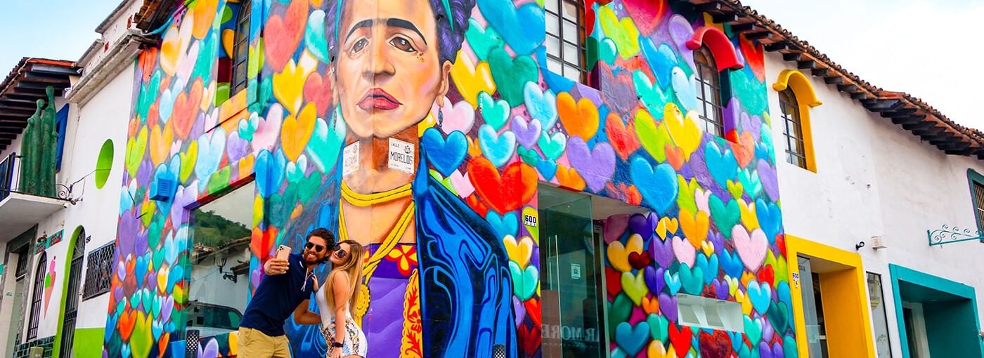 Couple taking a selfie in front of a colorful Frida Kahlo mural surrounded by multicolored hearts in Puerto Vallarta’s Historic Center.