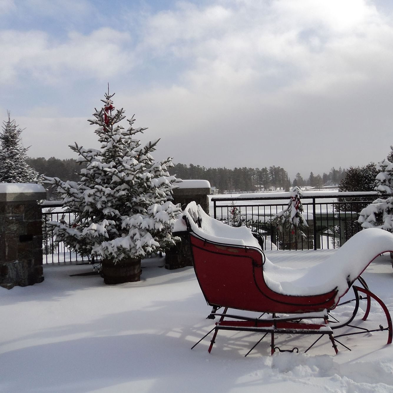 Snowy scene with a red sleigh and Christmas trees on a terrace.