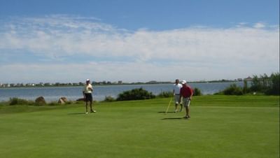 Three golfers by a lake as one lines up a shot under a clear blue sky near The Hotel Maria