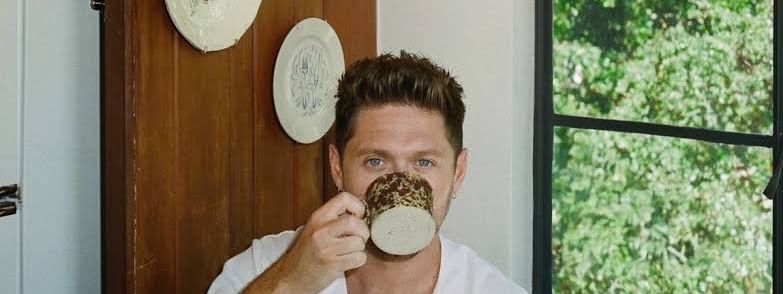 Man in white robe drinking from a decorative mug in a cozy room with wood paneling and decorative plates on the wall.